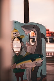 A colorful cartoon standee with cut-out face holes for photography features a person in a hat labeled 'Newport Landing' holding a large fish with a shark in the background. The scene seems humorous and playful, set against a backdrop of blurred outdoor elements.