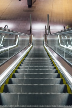 An empty escalator descends with silver steps and yellow safety lines. Glass railings with illuminated handrails run alongside, reflecting light. The floor at the bottom is visible in a muted color, lacking people or activity.