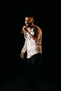 A man confidently sings into a microphone, dressed in a light-colored shirt with patterned sleeves and dark pants. He stands against a dark background that highlights his figure.