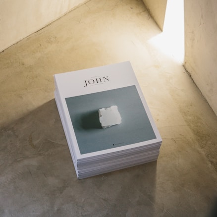 A stack of magazines titled 'JOHN' is placed on a smooth, concrete floor. The cover features a minimalist design with a white object in the center against a muted blue background. The image is illuminated by natural light coming from the top right corner.
