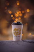 A takeaway coffee cup with a lid from Metro Café stands on a surface, against a blurred background of warm, glowing bokeh lights.