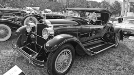 A classic vintage car with a sleek black finish and distinctive features such as large round headlights and intricate wire-spoke wheels. The car is parked on grass in a setting that suggests a car show, with other vintage vehicles visible in the background.