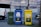 Three recycling bins of different colors stand side by side on a cobblestone street. The bins are labeled with the text 'Porto' and are designated for different types of waste: green for general waste, blue for paper, and yellow for plastics and metals. In the background, there is a vehicle and a building covered with scaffolding.