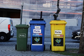 Three recycling bins of different colors stand side by side on a cobblestone street. The bins are labeled with the text 'Porto' and are designated for different types of waste: green for general waste, blue for paper, and yellow for plastics and metals. In the background, there is a vehicle and a building covered with scaffolding.