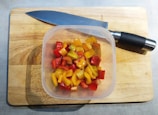 A plastic container filled with chopped red, yellow, and orange peppers is placed on a wooden cutting board. A large kitchen knife with a black handle rests nearby.
