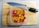 A plastic container filled with chopped red, yellow, and orange peppers is placed on a wooden cutting board. A large kitchen knife with a black handle rests nearby.