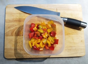 A plastic container filled with chopped red, yellow, and orange peppers is placed on a wooden cutting board. A large kitchen knife with a black handle rests nearby.