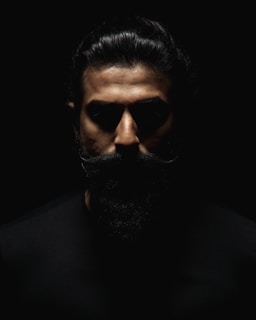 A dramatic portrait featuring a man with a prominent beard and slicked-back hair, emerging from deep shadows with intense lighting focused on the face.