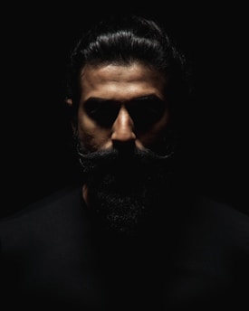 A dramatic portrait featuring a man with a prominent beard and slicked-back hair, emerging from deep shadows with intense lighting focused on the face.
