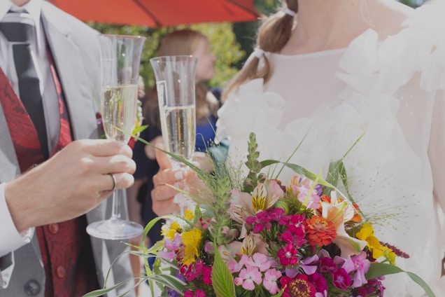 A couple is holding champagne flutes, toasting, with a colorful bouquet of flowers in the foreground. The man's attire includes a gray suit with a red waistcoat, while the woman is in a white outfit adorned with floral details. An umbrella providing shade is visible in the background.
