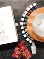 A collection of essential oil bottles are arranged in a semicircle on a wooden base next to a glass diffuser. There is a white box with branding and two red pouches labeled NingXia Red. A small bottle of hand purifier is placed nearby on a dark textured surface.