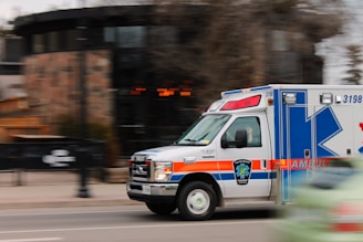 An ambulance is moving quickly down a street, captured with motion blur to emphasize speed. The vehicle is predominantly white with blue and orange markings, equipped with emergency lights on top. A blurred background indicates urban surroundings with some trees and buildings.