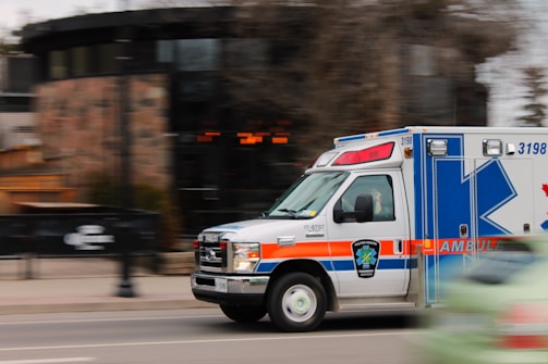 An ambulance is moving quickly down a street, captured with motion blur to emphasize speed. The vehicle is predominantly white with blue and orange markings, equipped with emergency lights on top. A blurred background indicates urban surroundings with some trees and buildings.