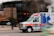An ambulance is moving quickly down a street, captured with motion blur to emphasize speed. The vehicle is predominantly white with blue and orange markings, equipped with emergency lights on top. A blurred background indicates urban surroundings with some trees and buildings.