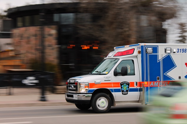 An ambulance is moving quickly down a street, captured with motion blur to emphasize speed. The vehicle is predominantly white with blue and orange markings, equipped with emergency lights on top. A blurred background indicates urban surroundings with some trees and buildings.