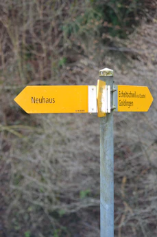 A metal post with two yellow directional signs pointing in different directions. The left sign reads 'Neuhaus' and the right sign shows 'Echeltschwil via Chastel, Goldingen.' The background consists of blurred trees and foliage.