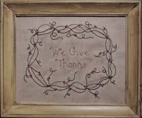 A piece of embroidery with the words 'We Give Thanks' surrounded by a decorative vine pattern that includes small flowers. Encased in a rustic wooden frame, the fabric appears to have a simple, natural texture.