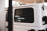 A close-up view of the rear side window of a white vehicle featuring a decal of a black and subdued American flag. The vehicle is parked possibly at a gas station, with part of a fuel pump visible in the foreground. The reflection on the window includes trees and a cloudy sky.