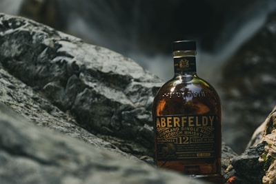 A bottle of Aberfeldy Highland Single Malt Scotch Whisky is placed outdoors on rocky terrain. The bottle is prominent in the foreground with its label clearly visible. The background features a blurred natural environment, suggesting a serene and rugged setting.