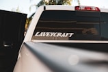 A close-up view of the rear window of a white vehicle, partially opened, with a large sticker saying 'LAVEYCRAFT'. The background shows blurred foliage and sky, and the sunlight reflects off the car's surface.