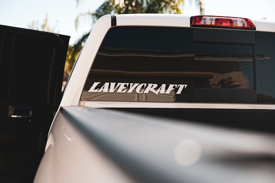 A close-up view of the rear window of a white vehicle, partially opened, with a large sticker saying 'LAVEYCRAFT'. The background shows blurred foliage and sky, and the sunlight reflects off the car's surface.