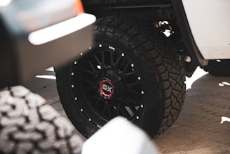 A close-up view of a vehicle's wheel featuring a black rim with a red and white center cap, surrounded by large, rugged off-road tires with noticeable tread patterns. The image captures part of the vehicle's white body and a shadowy, out-of-focus area in the foreground.