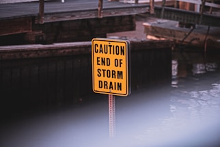 A bright yellow sign with black text reading 'CAUTION END OF STORM DRAIN' is positioned in front of a wooden structure near a body of water. The background is slightly out of focus.