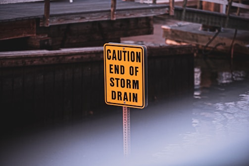 A bright yellow sign with black text reading 'CAUTION END OF STORM DRAIN' is positioned in front of a wooden structure near a body of water. The background is slightly out of focus.