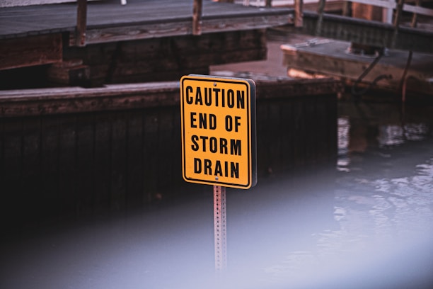 A bright yellow sign with black text reading 'CAUTION END OF STORM DRAIN' is positioned in front of a wooden structure near a body of water. The background is slightly out of focus.