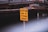 A bright yellow sign with black text reading 'CAUTION END OF STORM DRAIN' is positioned in front of a wooden structure near a body of water. The background is slightly out of focus.