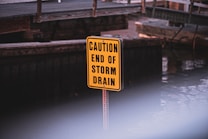 A bright yellow sign with black text reading 'CAUTION END OF STORM DRAIN' is positioned in front of a wooden structure near a body of water. The background is slightly out of focus.
