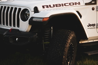 A close-up view of a white Jeep Wrangler Rubicon, highlighting the rugged front grille, branding, and heavily treaded tires. The vehicle is parked on grass, and the lighting suggests either early morning or late afternoon.