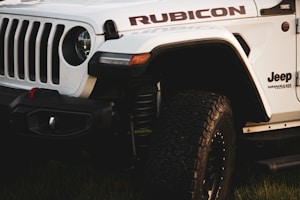 A close-up view of a white Jeep Wrangler Rubicon, highlighting the rugged front grille, branding, and heavily treaded tires. The vehicle is parked on grass, and the lighting suggests either early morning or late afternoon.