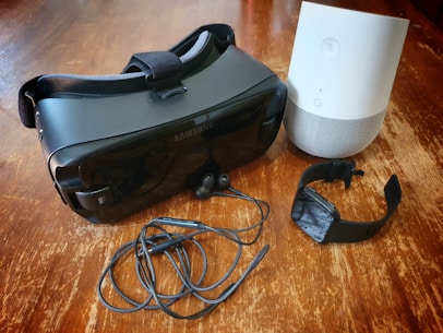 A collection of electronic gadgets arranged on a wooden surface, including a black virtual reality headset, a smart speaker in white and gray, black earbuds with a cable, and a black smartwatch.