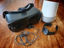 A collection of electronic gadgets arranged on a wooden surface, including a black virtual reality headset, a smart speaker in white and gray, black earbuds with a cable, and a black smartwatch.