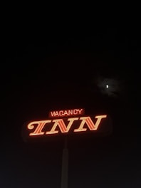 A neon sign displaying the word 'INN' prominently in bright red, with the word 'VACANCY' partially illuminated above it. The sign stands out against a dark nighttime sky, with the moon partially visible through some clouds.