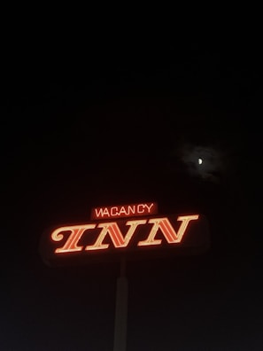 A neon sign displaying the word 'INN' prominently in bright red, with the word 'VACANCY' partially illuminated above it. The sign stands out against a dark nighttime sky, with the moon partially visible through some clouds.