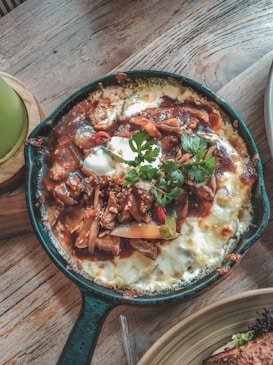 A cast iron skillet filled with a baked dish that includes melted cheese, a red sauce, garnished with green cilantro leaves, pieces of meat, sliced vegetables, and topped with a dollop of sour cream. The skillet is placed on a wooden surface with a partial view of a plate with bread on the side.