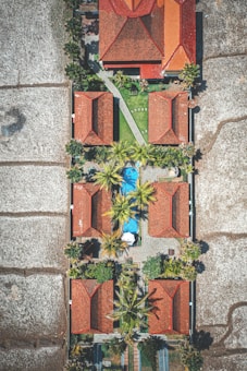 An aerial view of a symmetrical arrangement of buildings with red roofs surrounded by lush greenery and palm trees. A blue swimming pool is positioned centrally among the structures, and the entire area is enclosed by agricultural fields.