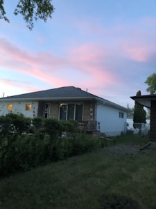 A suburban house with a stone and white exterior is surrounded by a well-maintained lawn and green shrubbery. The sky has a beautiful gradient of blue and pink hues, indicating either dawn or dusk. A tree branch is visible at the top left corner, adding to the natural ambiance.