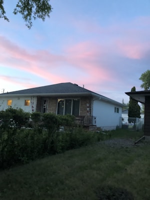 A suburban house with a stone and white exterior is surrounded by a well-maintained lawn and green shrubbery. The sky has a beautiful gradient of blue and pink hues, indicating either dawn or dusk. A tree branch is visible at the top left corner, adding to the natural ambiance.