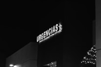 A black and white image of a building facade with the illuminated word 'URGENCIAS,' likely indicating an emergency entrance. The letters are adorned with small lights, and there is minimal visible street lighting.
