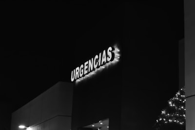 A black and white image of a building facade with the illuminated word 'URGENCIAS,' likely indicating an emergency entrance. The letters are adorned with small lights, and there is minimal visible street lighting.