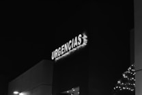 A black and white image of a building facade with the illuminated word 'URGENCIAS,' likely indicating an emergency entrance. The letters are adorned with small lights, and there is minimal visible street lighting.