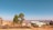A rural landscape features a cluster of simple structures with corrugated metal roofs and colorful paint. A few large trees stand prominently in the foreground, with rolling brown hills and clear blue sky stretching in the distance.