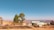 A rural landscape features a cluster of simple structures with corrugated metal roofs and colorful paint. A few large trees stand prominently in the foreground, with rolling brown hills and clear blue sky stretching in the distance.