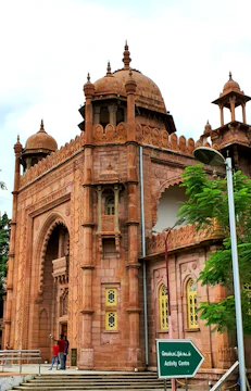 An ornate building with intricate architectural details, topped with domes and decorative spires. The facade features detailed carvings and arched windows, with latticed yellow window frames. A green sign indicates an activity center, while two people stand near the entrance next to a tree and a lamppost.