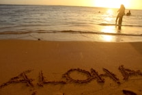 A tranquil beach scene with the word 'ALOHA' written in the sand. In the background, the sun is setting over the ocean, casting a warm golden glow on the water. People are visible near the shoreline, enjoying the evening light.