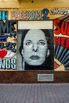 A wall features a mural with vibrant graffiti art. At the center is a monochrome portrait of a woman's face on a roller shutter. Surrounding the portrait are colorful words and designs, including the word 'freedom' painted in bold, diverse colors. The artwork juxtaposes words and images, creating a dynamic urban landscape.