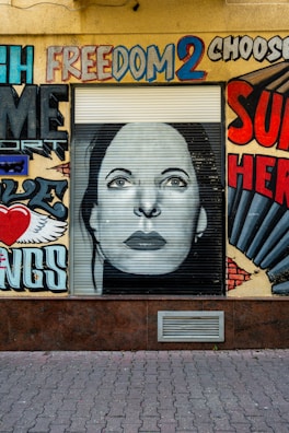 A wall features a mural with vibrant graffiti art. At the center is a monochrome portrait of a woman's face on a roller shutter. Surrounding the portrait are colorful words and designs, including the word 'freedom' painted in bold, diverse colors. The artwork juxtaposes words and images, creating a dynamic urban landscape.
